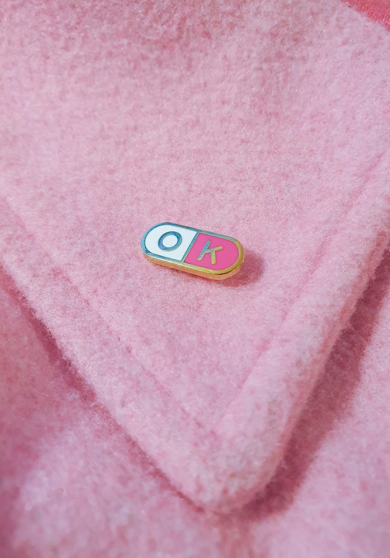 One of the enamel pins photographed on a light pink jacket coller. The pill shaped enamel pin is split in 2, with the wordK OK in the halves; O in the white half and K in the pink half.