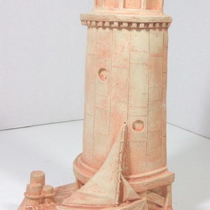 May include: A decorative lighthouse figurine in shades of peach and cream. The lighthouse features a detailed design with a small sailboat at its base. The figurine is made of a textured material, suggesting a weathered appearance.