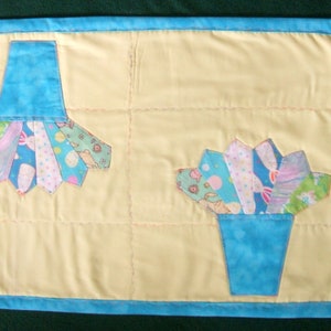 May include: A yellow quilted table runner with a blue border features two appliqued flower arrangements. Each arrangement has a blue pot and colorful fabric flowers.