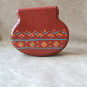 May include: A small, red, ceramic pot with a geometric pattern painted in orange and blue. The pot is shaped like a traditional Native American pot.