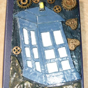 May include: A blue and white TARDIS, a time machine from the Doctor Who television series, is decorated with gold gears and clockwork embellishments. The TARDIS is set against a dark blue background.
