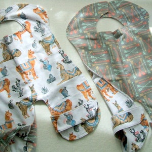 May include: Two baby bibs, one with a llama pattern and the other with an arrow pattern. Both bibs are made of soft, white fabric and have a neck opening.
