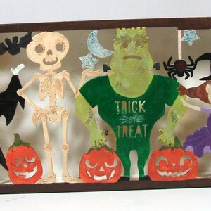 May include: A Halloween-themed papercraft scene with a vampire, skeleton, monster, and witch. The vampire is wearing a black cape and holding a red and white striped scarf. The skeleton is wearing a white shirt and black pants. The monster is wearing a green shirt with the words "Trick or Treat" on it. The witch is wearing a purple dress and a black hat. There are also pumpkins and a spider web in the scene.