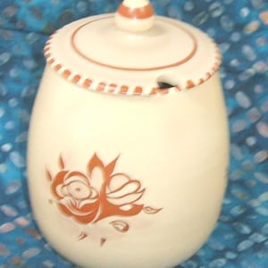 May include: Cream-colored ceramic sugar bowl with a lid and a floral design. The bowl has a rounded shape with a lid featuring a knob and a decorative rim. A floral motif in shades of brown adorns the front of the bowl. The sugar bowl is designed for kitchen use.