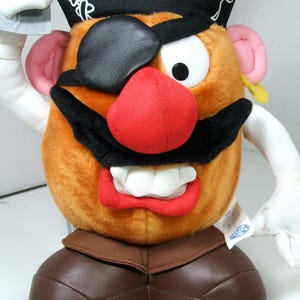May include: A plush toy of a pirate Mr. Potato Head with a black eyepatch, a red nose, a black mustache, and a white pirate hat with a skull and crossbones design. The toy is wearing brown pants and holding a sword.