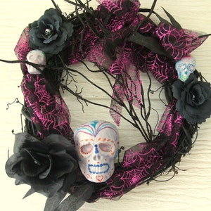 May include: A black wreath with pink and black spiderweb ribbon, black roses, and three sugar skulls. Two of the skulls are white with black details, and one is painted with colorful details.
