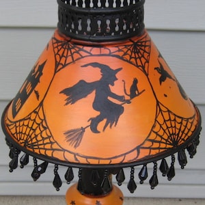 May include: A table lamp featuring an orange shade with black silhouettes of a witch on a broomstick, a cat, a haunted house, and spiderwebs. The lamp has a black metal base, a black beaded fringe, and a black stem with small orange star accents.