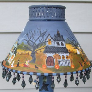 May include: A conical lamp shade featuring a hand-painted Halloween scene with a haunted house, bare trees, ghosts, and pumpkins. The shade has a dark blue perforated top rim and a bottom edge decorated with dangling dark grey teardrop beads.
