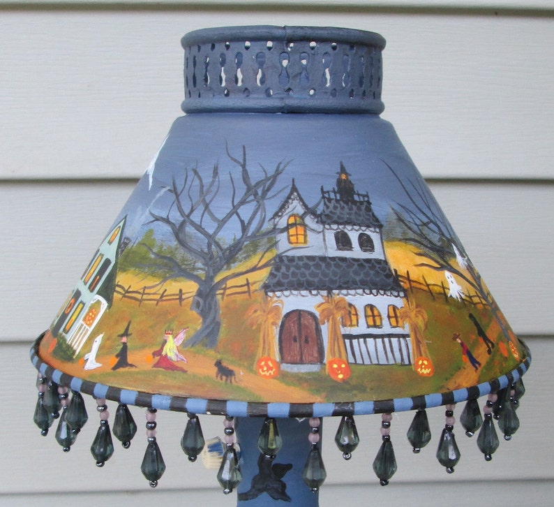 May include: A conical lamp shade featuring a hand-painted Halloween scene with a haunted house, bare trees, ghosts, and pumpkins. The shade has a dark blue perforated top rim and a bottom edge decorated with dangling dark grey teardrop beads.