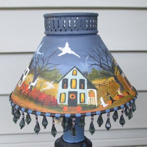 May include: A blue lampshade featuring a hand-painted Halloween scene with a house, ghosts, bare trees, and witches. The shade has a decorative metal top rim and a bottom edge adorned with dangling dark grey teardrop-shaped glass beads.