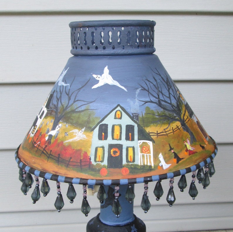 May include: A blue lampshade featuring a hand-painted Halloween scene with a house, ghosts, bare trees, and witches. The shade has a decorative metal top rim and a bottom edge adorned with dangling dark grey teardrop-shaped glass beads.