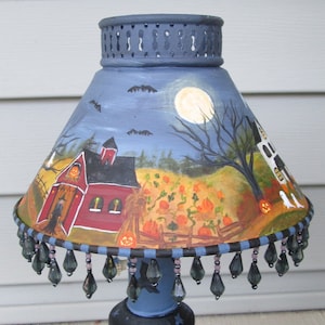 May include: A conical lamp shade featuring a hand-painted Halloween scene with a red barn, a pumpkin patch, a full moon, bats, and ghosts. The shade has a dark blue top rim and a bottom edge decorated with dangling dark grey teardrop-shaped beads.