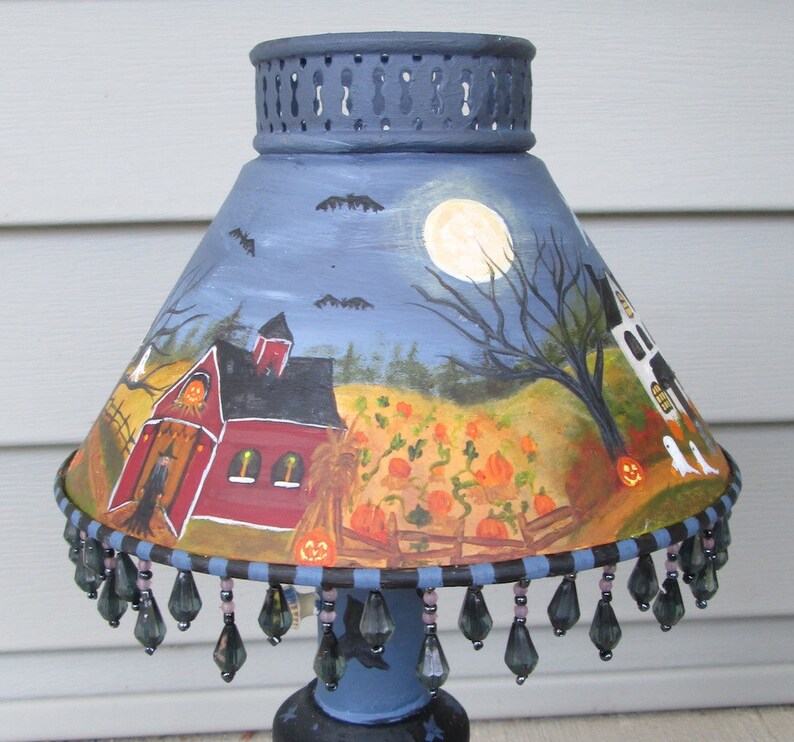 May include: A conical lamp shade featuring a hand-painted Halloween scene with a red barn, a pumpkin patch, a full moon, bats, and ghosts. The shade has a dark blue top rim and a bottom edge decorated with dangling dark grey teardrop-shaped beads.