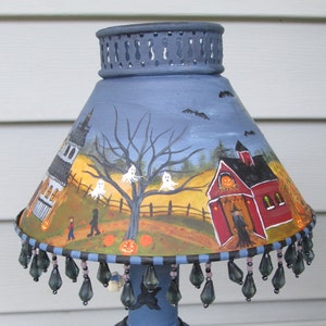 May include: A conical lamp shade painted with a Halloween scene featuring a red barn, a white house, ghosts, pumpkins, and a witch. The shade has a blue top rim with cutouts and a bottom edge decorated with dangling dark grey teardrop beads.