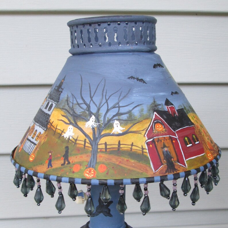 May include: A conical lamp shade painted with a Halloween scene featuring a red barn, a white house, ghosts, pumpkins, and a witch. The shade has a blue top rim with cutouts and a bottom edge decorated with dangling dark grey teardrop beads.