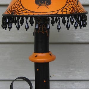 May include: A table lamp featuring an orange shade with black illustrations of ghosts, a cauldron, and spiderwebs. The base is black and orange with spider motifs. The shade is trimmed with dangling black teardrop-shaped beads and a black metal top.