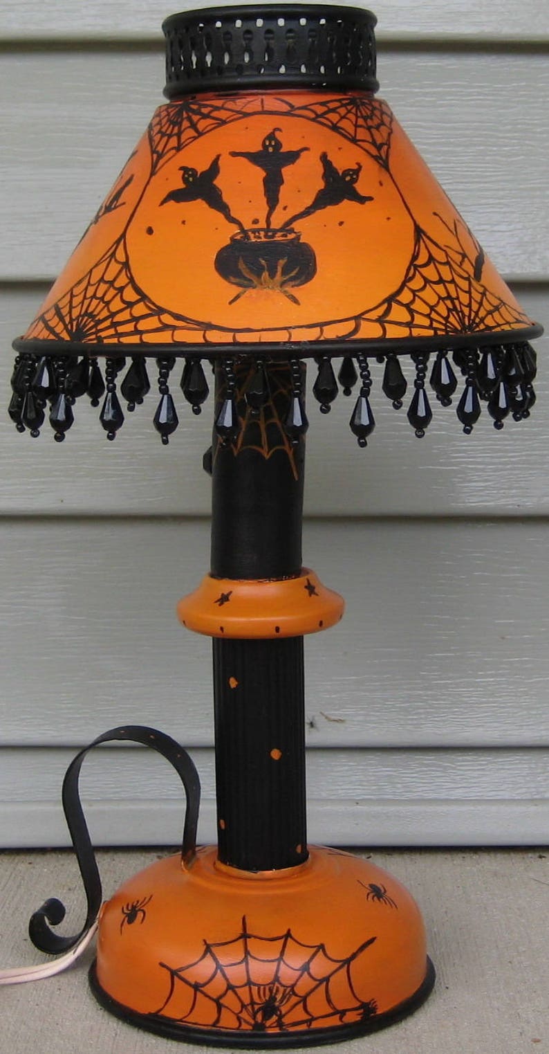 May include: A table lamp featuring an orange shade with black illustrations of ghosts, a cauldron, and spiderwebs. The base is black and orange with spider motifs. The shade is trimmed with dangling black teardrop-shaped beads and a black metal top.