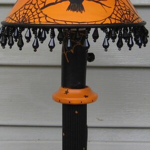 May include: A table lamp featuring an orange lampshade with a black owl and spiderweb design, accented by a black beaded fringe. The base is black and orange with painted spiderwebs and stars, supported by a round orange base with a spider graphic.