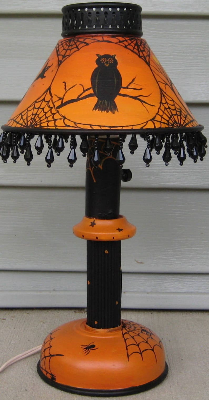 May include: A table lamp featuring an orange lampshade with a black owl and spiderweb design, accented by a black beaded fringe. The base is black and orange with painted spiderwebs and stars, supported by a round orange base with a spider graphic.