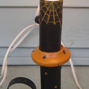 May include: A decorative table lamp featuring a black cylindrical stem with a yellow spiderweb design and an orange base painted with black spiders and webs. The lamp has a curved black handle and is topped with a glowing white light bulb.