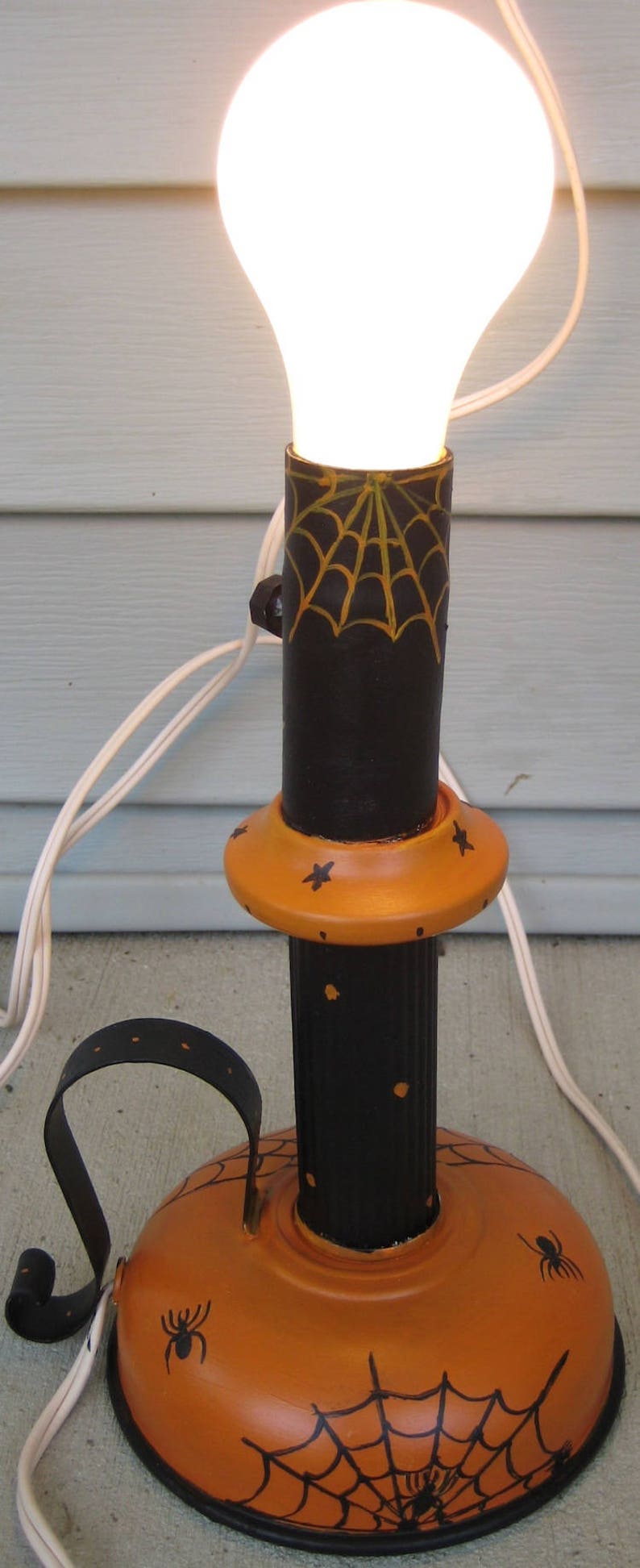 May include: A decorative table lamp featuring a black cylindrical stem with a yellow spiderweb design and an orange base painted with black spiders and webs. The lamp has a curved black handle and is topped with a glowing white light bulb.