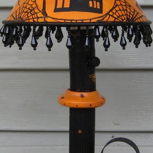 May include: A table lamp featuring an orange lampshade with black illustrations of a haunted house, spiderwebs, and bats. The shade has a black beaded fringe. The base is black and orange with spiderweb motifs and a curved metal handle.