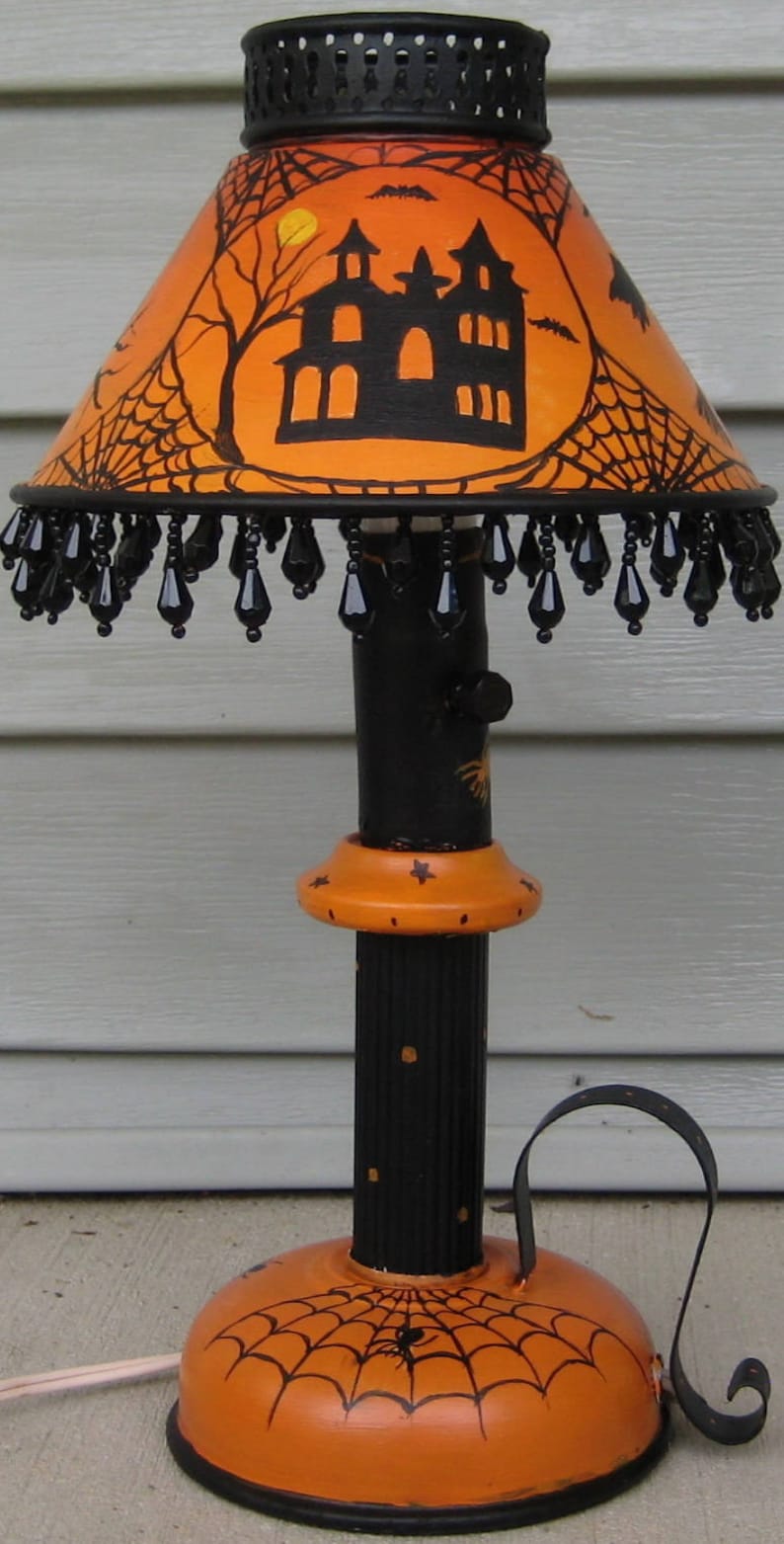 May include: A table lamp featuring an orange lampshade with black illustrations of a haunted house, spiderwebs, and bats. The shade has a black beaded fringe. The base is black and orange with spiderweb motifs and a curved metal handle.