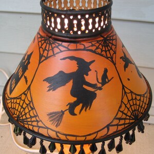 May include: A conical orange lampshade featuring black hand-painted silhouettes of a witch on a broomstick with a cat, a haunted house, and spiderwebs. The shade has a black metal top rim with cutouts and a bottom edge trimmed with black beads.