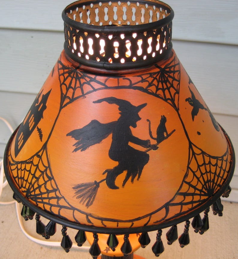 May include: A conical orange lampshade featuring black hand-painted silhouettes of a witch on a broomstick with a cat, a haunted house, and spiderwebs. The shade has a black metal top rim with cutouts and a bottom edge trimmed with black beads.