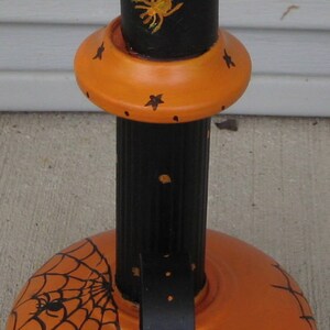 May include: A black and orange metal stand featuring a cylindrical post and a wide, rounded base. The black post is decorated with yellow spiderwebs and a spider, while the orange base displays black spiderwebs and spiders with small stars.