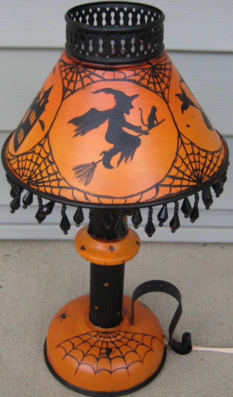 May include: A table lamp featuring an orange shade with black silhouettes of a witch on a broomstick, a cat, a haunted house, and spiderwebs. The lamp has a black metal base, a black beaded fringe, and a black stem with small orange star accents.