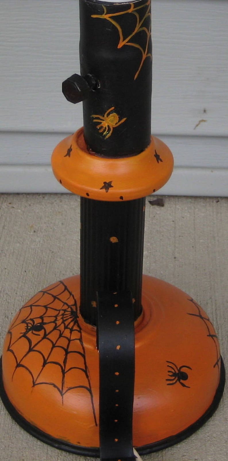 May include: A black and orange metal stand featuring a cylindrical post and a wide, rounded base. The black post is decorated with yellow spiderwebs and a spider, while the orange base displays black spiderwebs and spiders with small stars.