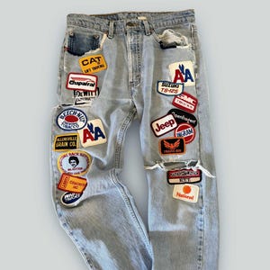 May include: Light blue denim jeans with multiple patches featuring logos and text. Patches include "CAT Lift Trucks", "Jeep", "Suzuki TS-125", and "Freightliner E.T.T.". The jeans have distressed details and ripped knees.