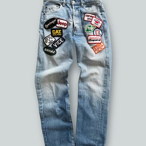 May include: Light blue denim jeans with various embroidered patches. Patches include the words "Copenhagen", "Jeep", "CAT Diesel Power", "Indy", "Vice", "Skoal", "Four Wheel Drive", "Pele", "Chaparral", and "Etown Distributing Co. Inc."