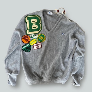 May include: A gray V-neck sweater adorned with various patches. The sweater features a large green and white "B" patch, along with smaller patches displaying text and graphics. A small blue logo is visible on the chest.