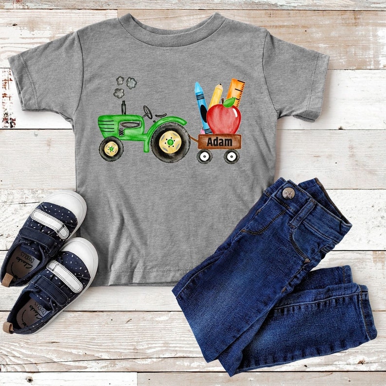 May include: A green toy tractor pulling a wagon with school supplies, including a red apple, crayons, and a ruler. The wagon is labeled with the name "Adam".