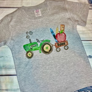 May include: A gray t-shirt with a green tractor pulling a wagon full of school supplies, including a red apple, pencils, and a ruler. The wagon says "Rylee" on the side.