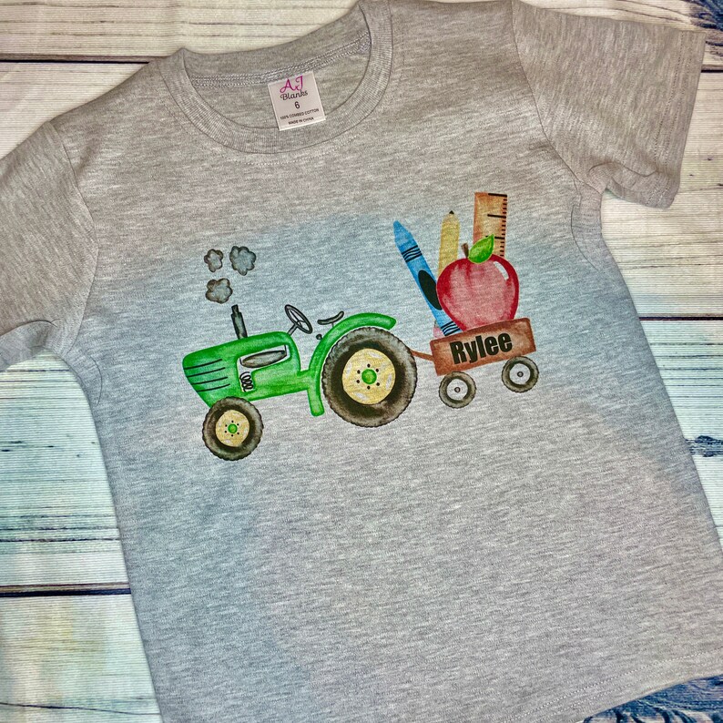 May include: A gray t-shirt with a green tractor pulling a wagon full of school supplies, including a red apple, pencils, and a ruler. The wagon says "Rylee" on the side.