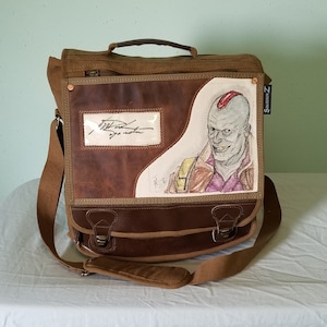 May include: A brown canvas and leather messenger bag with a hand-drawn illustration of a character on the front. The bag has a top handle, a shoulder strap, and two front buckles. The illustration features a figure with a red mohawk and a purple shirt.