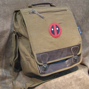 May include: A tan canvas messenger bag with a brown leather flap and accents. The bag features a Deadpool logo patch and a shoulder strap. The bag has a zipper closure and metal clasps.