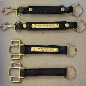 May include: Three leather keychains with brass hardware. The top keychain has a plain leather strap with a brass buckle. The middle keychain has a leather strap with a brass plate that reads "2024 AMHR Nationals". The bottom keychain has a leather strap with a brass plate that reads "AMHR Nationals Top Ten".