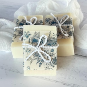 May include: Four bars of soap are wrapped in floral patterned paper and tied with white twine. The paper features a blue and cream floral design. The soap bars are a light cream color and rest on a white surface.