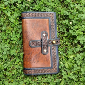 May include: A brown leather journal with a blue border and a decorative strap with brass accents. The journal is lying on a bed of green clover.