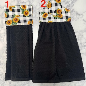 May include: Two black kitchen towels with a sunflower and black and white checkered pattern at the top. One towel is folded, and the other is hanging. The towels are made of a textured fabric.