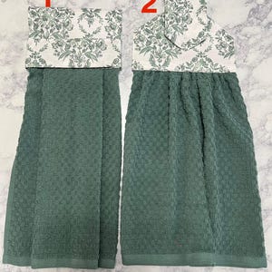 May include: Two hanging kitchen towels. The towels are a solid teal green color with a textured weave. The top portion of each towel features a white fabric with a green floral pattern. The towels are designed to hang over an oven or cabinet handle.