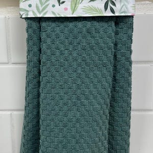 May include: A dark teal kitchen towel with a textured waffle weave pattern. The top section features a white fabric band decorated with a pattern of green leaves and small pink dots, folded over a silver metal bar for hanging display.