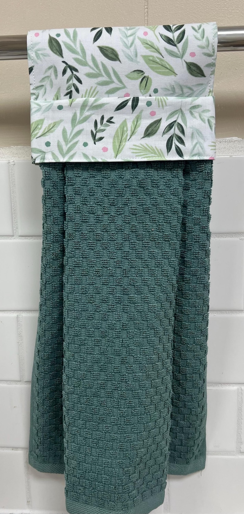 May include: A dark teal kitchen towel with a textured waffle weave pattern. The top section features a white fabric band decorated with a pattern of green leaves and small pink dots, folded over a silver metal bar for hanging display.