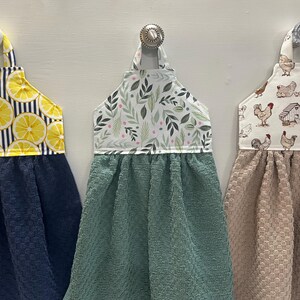 May include: Three hanging kitchen towels with fabric tops and textured bottoms. The left towel has a yellow lemon and navy stripe top with a navy bottom. The center towel has a green leaf print top with a sage green bottom. The right towel has a chicken print top with a tan bottom.