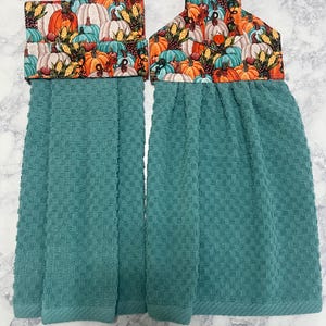 May include: Two hanging kitchen towels featuring a textured teal waffle-weave fabric. The top sections are made of a fabric printed with a colorful pattern of pumpkins, corn, and berries in shades of orange, white, and blue, secured with a button.