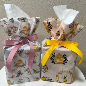 May include: Two square tissue box covers made of fabric featuring a bee and beehive pattern. The left cover is white with a pink ribbon tied in a bow, while the right cover is light peach with a bright yellow ribbon tied in a bow at the top.